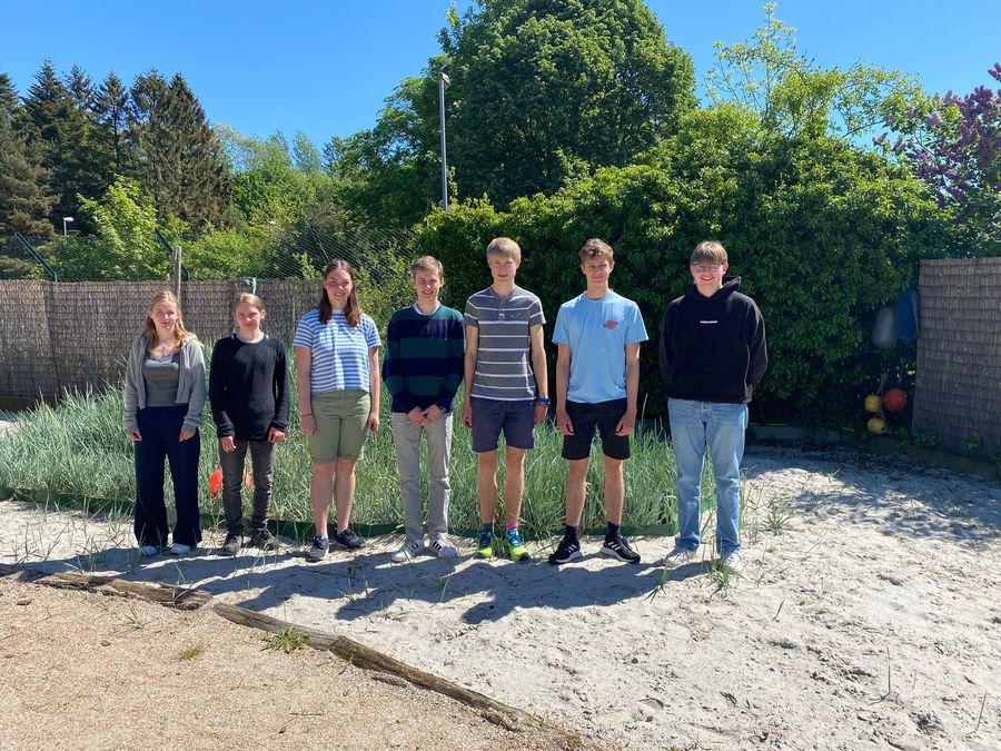 Die sieben schleswig-holsteinischen Landessiegerinnen und -sieger des Wettbewerbs Jugend forscht stehen im Strandbereich der Kieler Forschungswerkstatt vor einer künstlich angelegten kleinen Düne.